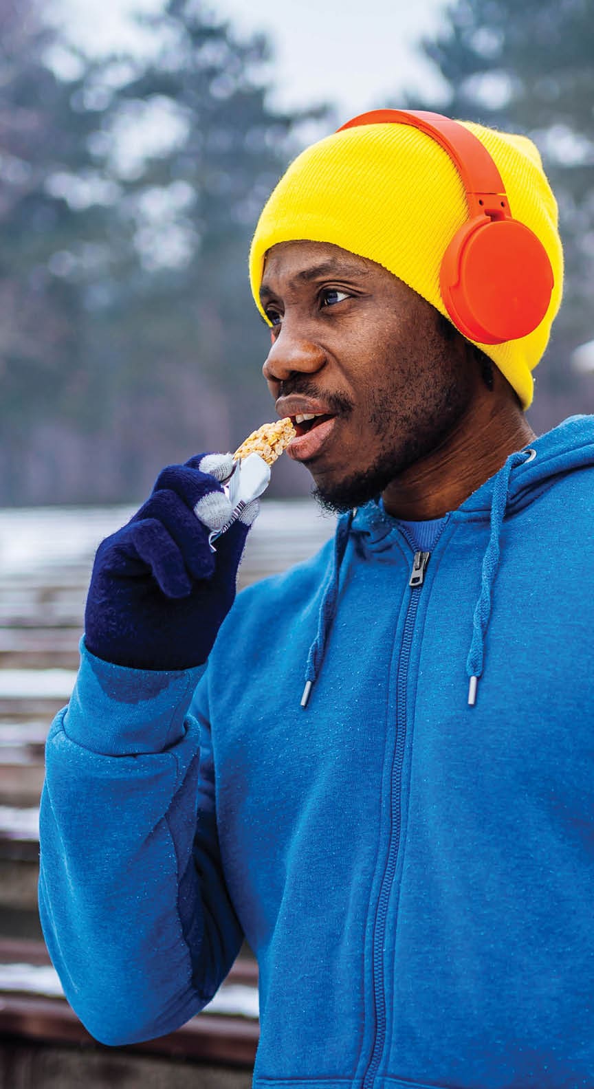 Young cheerful sporty man taking a break and eating a cereal bar during running pause on cold winter day